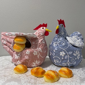 May include: Two fabric chicken-shaped bread warmers, one pink and one blue, with floral patterns. The pink warmer holds several sesame seed buns. The chickens have red combs and yellow beaks. Several sesame seed buns are on the table.