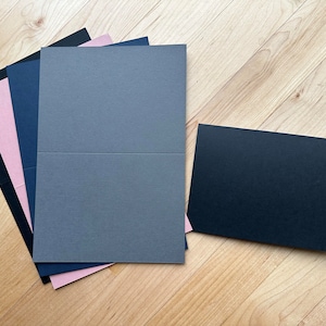May include: A stack of four cardstock sheets in different colors: black, pink, blue, and gray. The gray sheet is folded in half. A separate black cardstock sheet is lying on the wooden surface.