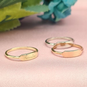 Può includere: Tre anelli d'oro con una fascia piatta rettangolare. L'anello superiore ha il nome "Caroline" inciso sulla fascia. Gli anelli sono su una superficie rosa.