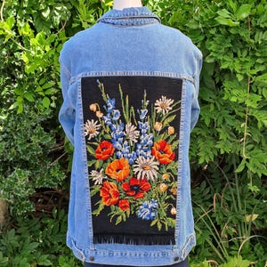 May include: A light blue denim jacket featuring a large, rectangular black fabric panel sewn onto the back. The panel displays a vibrant, detailed needlepoint embroidery of various wildflowers, including red poppies, blue delphiniums, and white daisies.