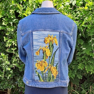May include: A blue denim jacket featuring a rectangular needlepoint panel on the back. The panel displays a detailed embroidered design of yellow iris flowers with green stems and leaves set against a light blue, horizontally striped background.