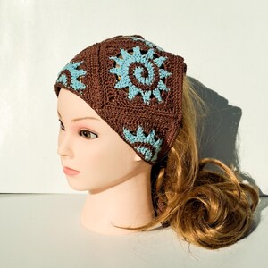 Könnte beinhalten: Ein braunes Häkelstirnband mit einem quadratischen Muster mit hellblauen floralen Designs. Das Stirnband wird auf einem Schaufensterkopf mit langen, hellbraunen Haaren getragen. Ein stilvolles Accessoire.