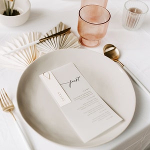 May include: A white place setting with a white plate, gold cutlery, and a white menu card with the word "feast" written in black calligraphy. The menu card is labelled "Andre" and includes a list of food items. The table is decorated with white paper fans and pink glasses.