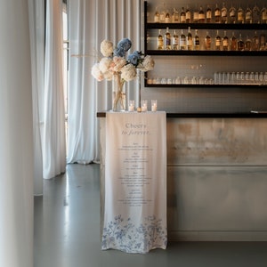 May include: A white linen tablecloth with blue floral print and the text "Cheers to forever..." hangs on a bar counter. The text lists various drinks, including margaritas, beer, wine, and spirits. A bouquet of white and blue flowers sits in a clear vase on the counter.