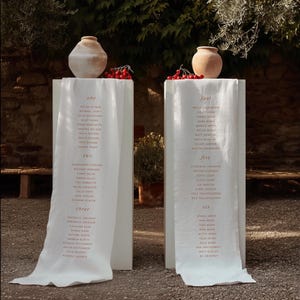 Pode incluir: Dois caminhos de mesa de tecido branco drapeados sobre pedestais retangulares altos, cada um coroado com um vaso de cerâmica bege e bagas vermelhas. Os caminhos apresentam listas de nomes impressas sob os cabeçalhos um, dois, três, quatro, cinco e seis.