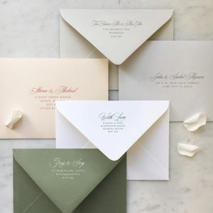 May include: A collection of five rectangular envelopes in cream, light grey, pale peach, white, and sage green. Each envelope features elegant script and serif typography displaying names and addresses, accompanied by three small white flower petals.