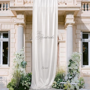 Può includere: Un lungo striscione verticale in tessuto di raso bianco sporco appeso a un balcone. Lo striscione presenta il testo nero "The Hendricks" in corsivo, seguito dal testo più piccolo "Est. 2026" stampato vicino alla parte inferiore centrale.