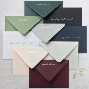 Pode incluir: Uma variedade de envelopes coloridos em v&aacute;rios tons, incluindo verde, preto e bord&ocirc;. Cada envelope apresenta texto dourado que diz "with love" ou "kindly deliver to". Os envelopes est&atilde;o dispostos sobre uma superf&iacute;cie branca.