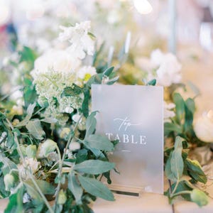 May include: A frosted acrylic table sign with the words "Top TABLE" in elegant white script. The sign is surrounded by lush greenery and white flowers, creating a soft, romantic aesthetic, perfect for a wedding or event.