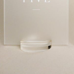 May include: A clear, curved acrylic display stand holding a white card with the word "FIVE" at the top. The stand is positioned at the bottom of the card, creating a minimalist aesthetic.
