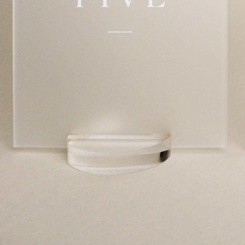 May include: A clear, curved acrylic display stand holding a white card with the word "FIVE" at the top. The stand is positioned at the bottom of the card, creating a minimalist aesthetic.