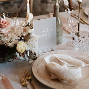 May include: A table setting with a white card that reads "Table ONE" in elegant script. The table is set with gold-rimmed glassware, a white plate, a folded napkin, and gold forks. Floral arrangements and a lit candle add to the ambiance.