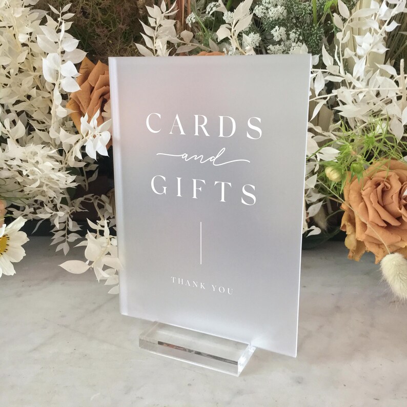 May include: A frosted acrylic sign with the words "CARDS and GIFTS" in white script and block letters. The sign includes a vertical line and the words "THANK YOU" at the bottom. It is displayed on a clear acrylic base, with floral arrangements in the background.