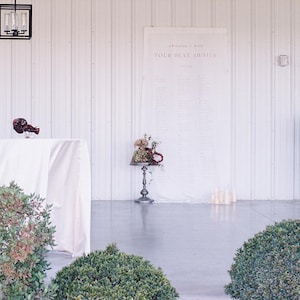 Puede incluir: Escena de recepción de boda con una mesa blanca con un arreglo floral, un letrero con el texto "YOUR SEAT AWAITS" y un soporte decorativo de metal con flores. La escena está ambientada en un espacio luminoso y abierto con arbustos verdes.