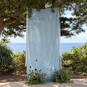 May include: A light blue fabric wedding seating chart banner with the text "YOUR SEAT AWAITS" printed at the top, featuring columns of guest names, white lace trim on the sides, and arrangements of sunflowers and blue thistles at the base.