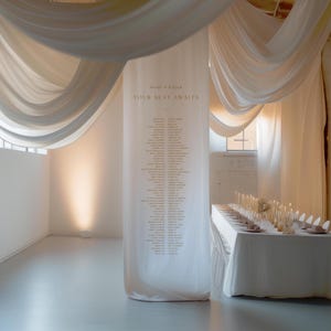 Puede incluir: Un telón de fondo de tela blanca con el texto "Rise & Edgar Your Seat Awaits" impreso en él. Una larga mesa blanca con velas y cubiertos está preparada frente al telón de fondo.