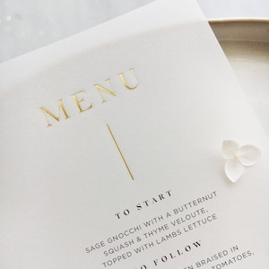 Può includere: Primo piano di un menu bianco con scritte dorate. La parola "MENU" è in alto, con una linea verticale sotto. Il menu elenca piatti come gnocchi alla salvia. Un piccolo fiore bianco è nell'angolo.