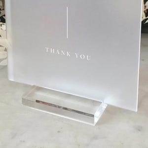 May include: A frosted white sign with the words "THANK YOU" in a simple font. A vertical line is above the text. The sign is held in a clear acrylic stand. The overall design is minimalist and elegant.