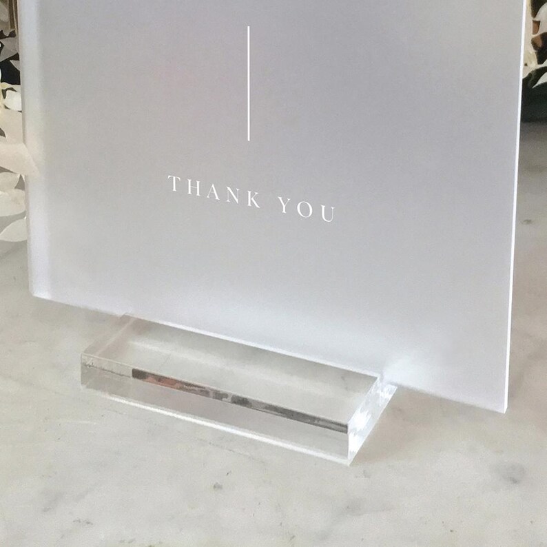 May include: A frosted white sign with the words "THANK YOU" in a simple font. A vertical line is above the text. The sign is held in a clear acrylic stand. The overall design is minimalist and elegant.