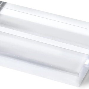 May include: A clear acrylic block with a rectangular shape. The block has a slot cut into the top, creating a space for holding items. The edges are smooth and the material is transparent, allowing light to pass through.