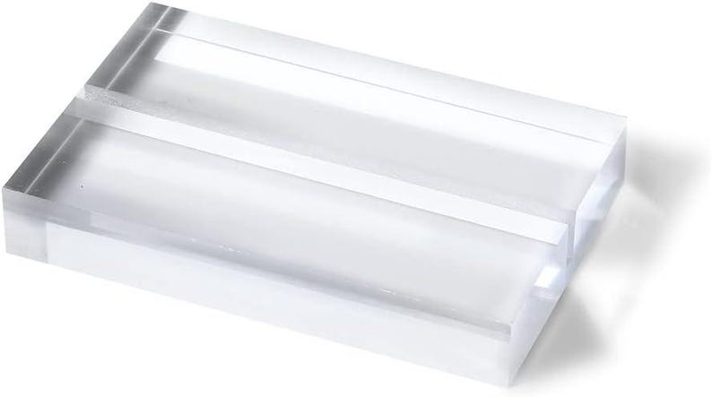 May include: A clear acrylic block with a rectangular shape. The block has a slot cut into the top, creating a space for holding items. The edges are smooth and the material is transparent, allowing light to pass through.