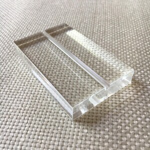 May include: A clear acrylic table number holder with a rectangular shape. The holder has a slot in the middle to hold a card. The edges are smooth and polished. The holder is sitting on a textured, off-white surface.