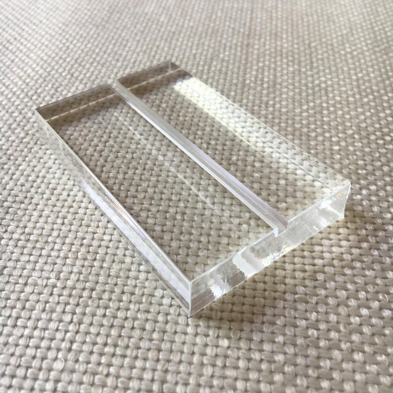 May include: A clear acrylic table number holder with a rectangular shape. The holder has a slot in the middle to hold a card. The edges are smooth and polished. The holder is sitting on a textured, off-white surface.