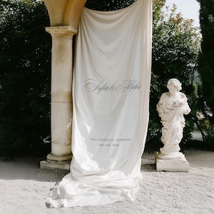 May include: A large, off-white fabric backdrop featuring the printed text Sofia & Miles, VILLA POZZOLO, FLORENCE, and June 6th, 2026. The material hangs vertically with soft folds, draping onto the ground in a smooth, flowing arrangement.