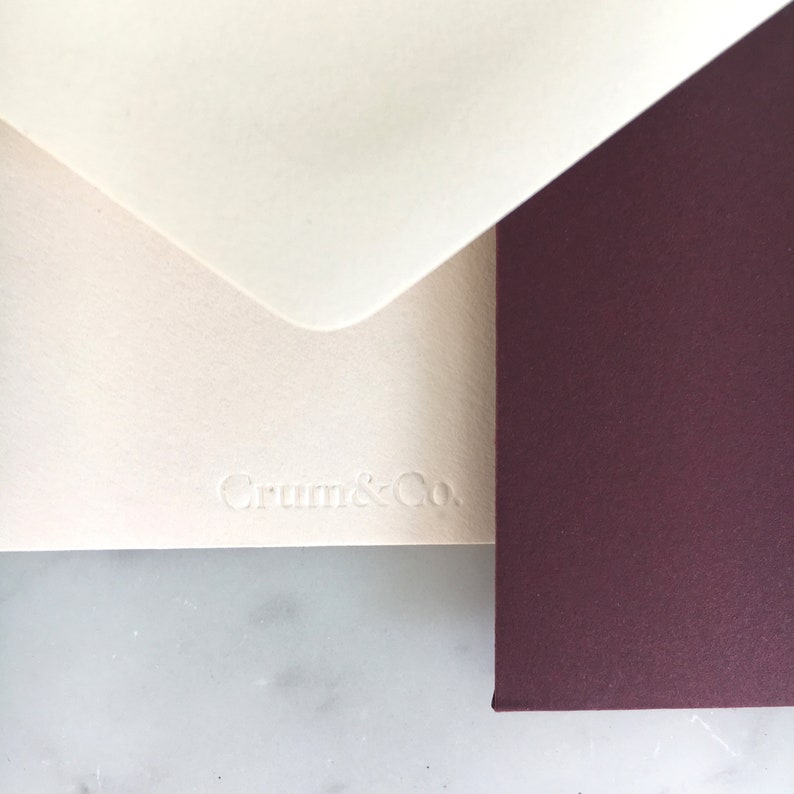Pode incluir: Close-up de artigos de papelaria. Um envelope creme com o texto em relevo "Crum & Co." &eacute; parcialmente vis&iacute;vel, juntamente com um cart&atilde;o bord&ocirc; escuro. O envelope &eacute; sobreposto com uma aba branca.