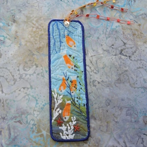 May include: A rectangular fabric bookmark featuring an underwater scene with orange fish, white coral, and green seaweed embroidered on a blue quilted background. The bookmark is finished with a dark blue border and a decorative orange beaded tassel.