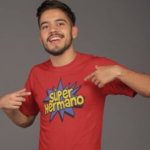 May include: Red t-shirt with the text "Super Hermano" in yellow and blue, a comic book style design. The shirt is paired with blue jeans.