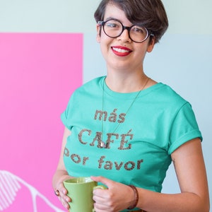May include: A woman wearing a turquoise t-shirt with the text "más café por favor" in brown letters, holding a green mug. She is smiling and wearing glasses.