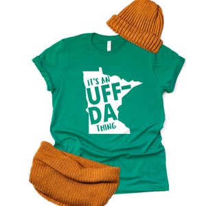 Könnte beinhalten: Ein grünes T-Shirt mit einer weißen Umrandung des Bundesstaates Minnesota und dem Text "It's an Uff-Da Thing". Das Shirt wird mit einer braunen Strickmütze und einem braunen Strickschal kombiniert.