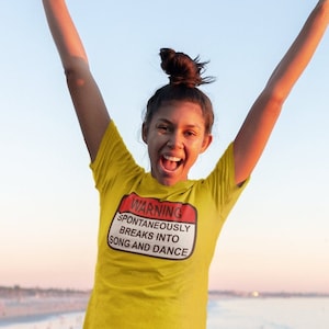 Puede incluir: Una mujer con una camiseta amarilla con el texto "WARNING SPONTANEOUSLY BREAKS INTO SONG AND DANCE" salta en el aire en un muelle con vistas al océano.