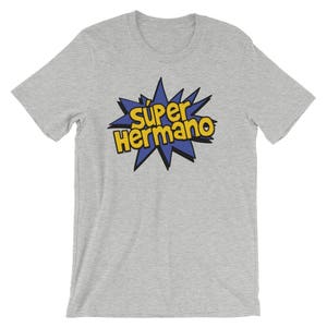 May include: A light gray t-shirt featuring the text "Super Hermano" in yellow and black, set against a blue starburst design. The shirt is made of a soft material and has short sleeves.