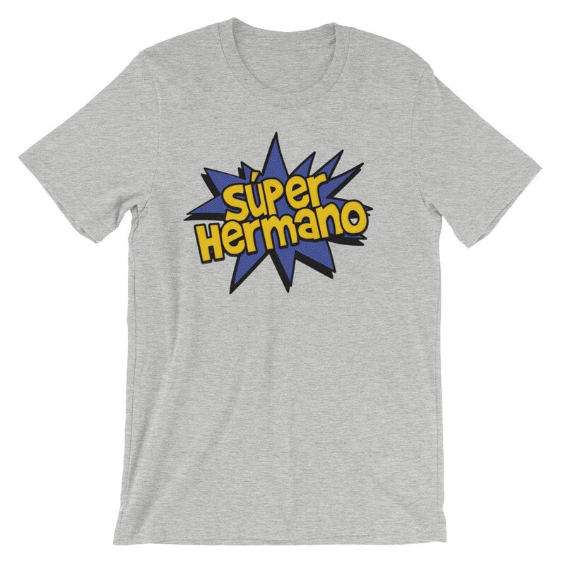 May include: A light gray t-shirt featuring the text "Super Hermano" in yellow and black, set against a blue starburst design. The shirt is made of a soft material and has short sleeves.