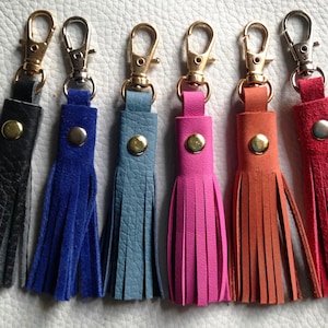 Puede incluir: Seis llaveros de cuero con borlas en varios colores, incluyendo negro, azul, azul claro, rosa, naranja y rojo. Cada llavero tiene un cierre de metal dorado o plateado.