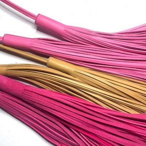 Puede incluir: Cuatro borlas de cuero en colores rosa y marrón. Las borlas están hechas de tiras finas de cuero que están atadas juntas en la parte superior. Las borlas cuelgan de un fondo blanco.