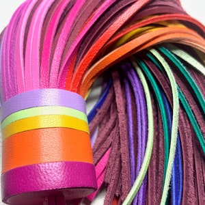 Op de afbeelding: Een kleurrijke leren kwast met een regenboog aan kleuren, waaronder roze, rood, oranje, geel, groen, blauw en paars. De kwast is bevestigd aan een dik, veelkleurig leren koord.