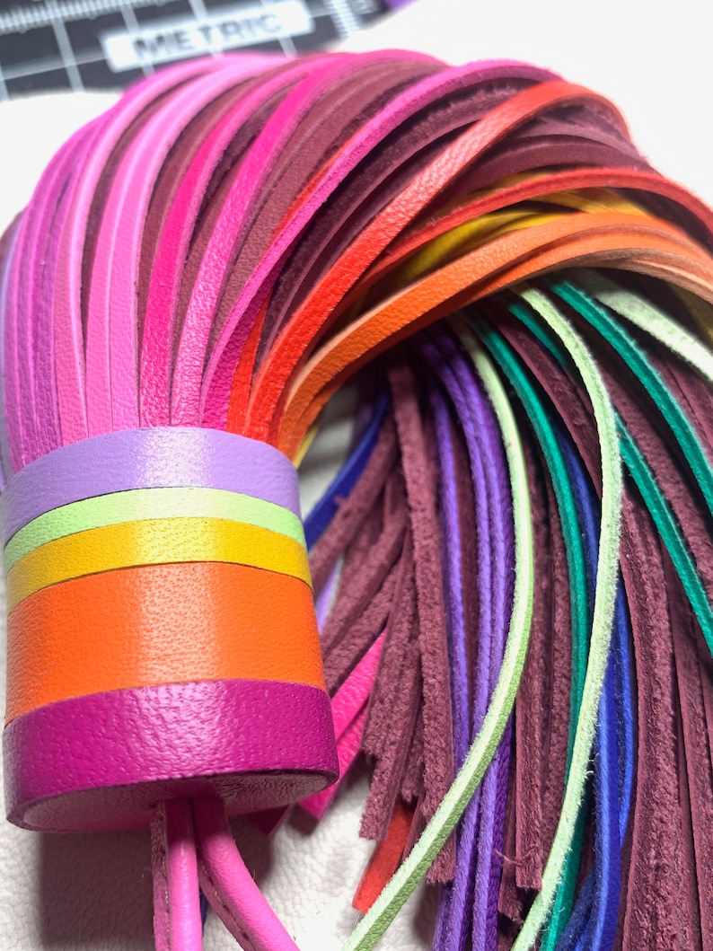 Op de afbeelding: Een kleurrijke leren kwast met een regenboog aan kleuren, waaronder roze, rood, oranje, geel, groen, blauw en paars. De kwast is bevestigd aan een dik, veelkleurig leren koord.