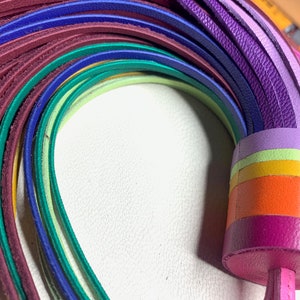 Op de afbeelding: Een regenboogkleurig leren koord met een kwast aan het uiteinde. Het koord is gemaakt van meerdere strengen leer in verschillende kleuren, waaronder rood, oranje, geel, groen, blauw en paars.