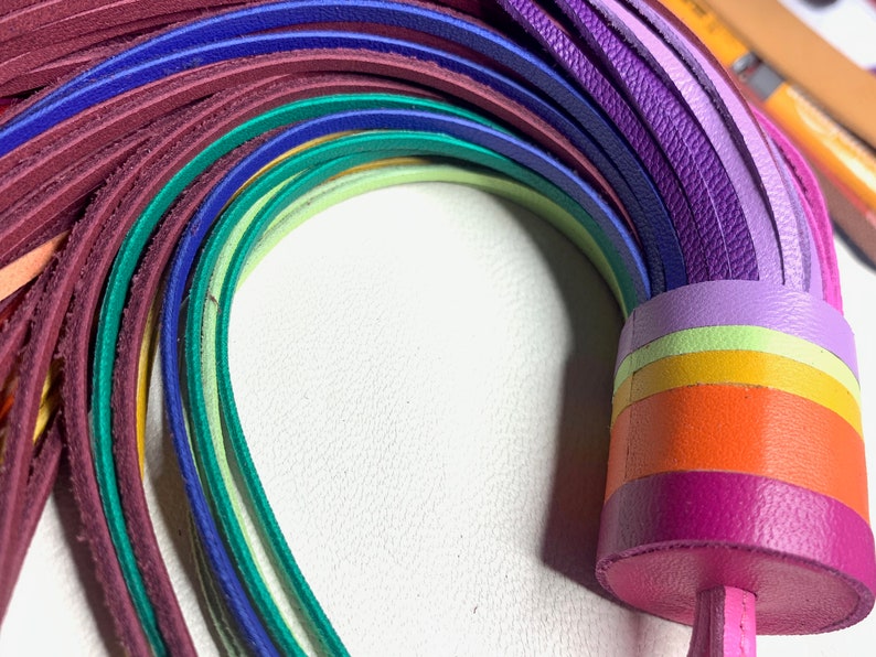 Op de afbeelding: Een regenboogkleurig leren koord met een kwast aan het uiteinde. Het koord is gemaakt van meerdere strengen leer in verschillende kleuren, waaronder rood, oranje, geel, groen, blauw en paars.
