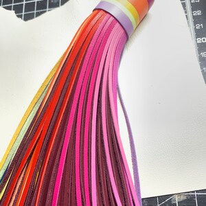 Op de afbeelding: Een kleurrijke leren kwast sleutelhanger met een regenboog van kleuren, waaronder rood, oranje, geel, groen, blauw, paars en roze. De kwast is bevestigd aan een paarse leren band met een metalen ring.