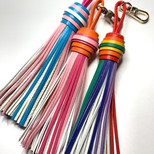 Könnte beinhalten: Drei farbenfrohe Schlüsselanhänger mit Quasten aus Leder. Die Quasten sind rosa, blau, orange, grün, lila und rot. Jeder Schlüsselanhänger hat einen goldfarbenen Verschluss und eine passende Lederschlaufe. Die Quasten bestehen aus dünnen Lederstreifen.
