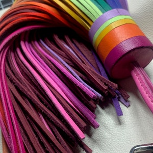Op de afbeelding: Een regenboogkleurige leren kwast sleutelhanger met een roze leren band. De kwast is gemaakt van meerdere strengen leer in verschillende tinten rood, oranje, geel, groen, blauw, paars en roze.