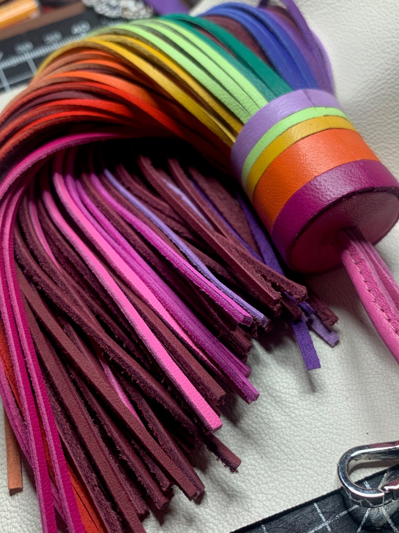 Op de afbeelding: Een regenboogkleurige leren kwast sleutelhanger met een roze leren band. De kwast is gemaakt van meerdere strengen leer in verschillende tinten rood, oranje, geel, groen, blauw, paars en roze.