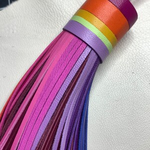 Op de afbeelding: Een veelkleurige leren kwast met een regenboog van kleuren, waaronder paars, roze, oranje, geel en groen. De kwast is gemaakt van dunne leren stroken die aan de bovenkant aan elkaar zijn gebonden.