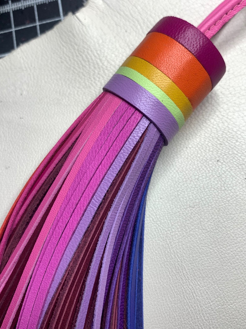 Op de afbeelding: Een veelkleurige leren kwast met een regenboog van kleuren, waaronder paars, roze, oranje, geel en groen. De kwast is gemaakt van dunne leren stroken die aan de bovenkant aan elkaar zijn gebonden.