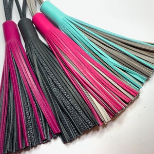 May include: Four leather tassels in different colors: pink, black, gray, and turquoise. The tassels are made of multiple strands of leather and have a fringe at the bottom.