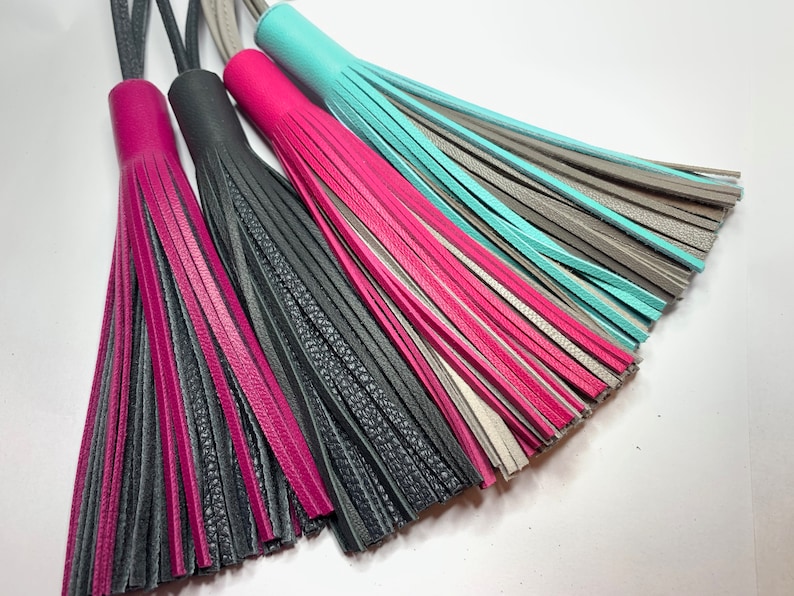 May include: Four leather tassels in different colors: pink, black, gray, and turquoise. The tassels are made of multiple strands of leather and have a fringe at the bottom.